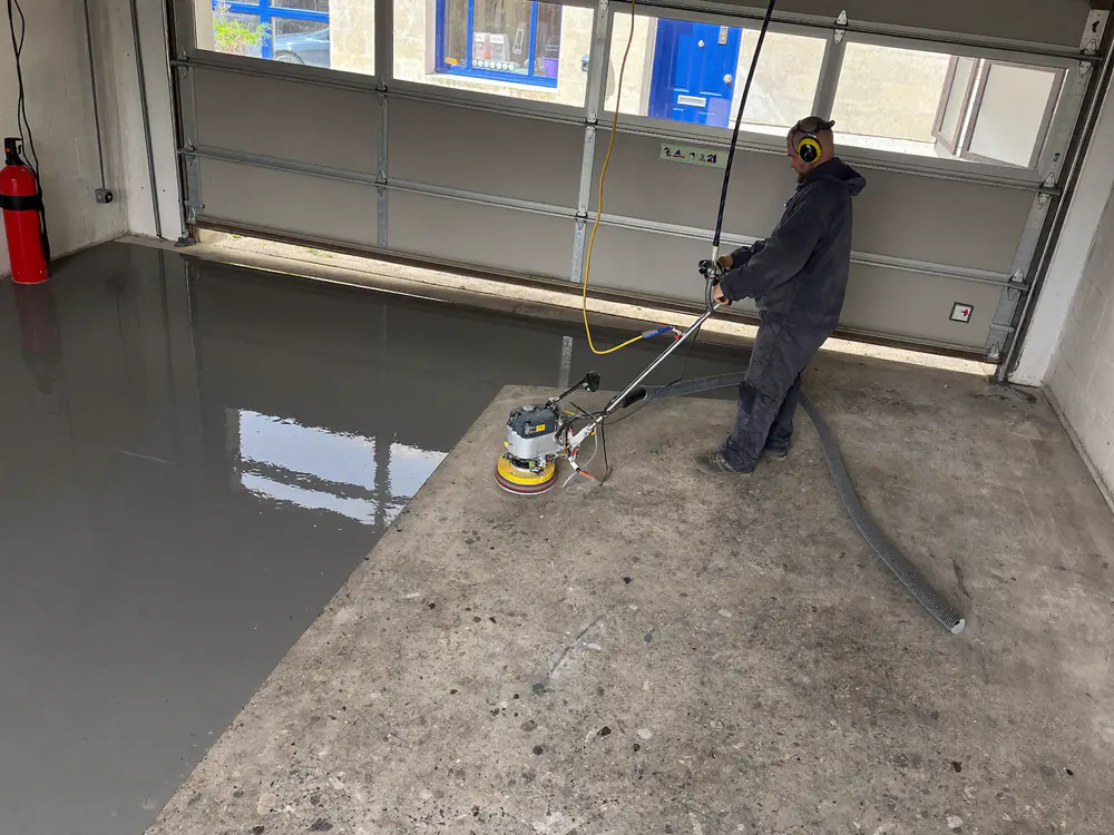 Een vloerslijper met stofafzuiging slijpt een grijze epoxycoating van een betonvloer in een garage