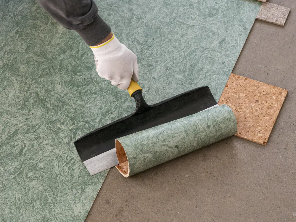 Een groen gemarmerd linoleum wordt met een brede schraper losgestoken, met bruine lijm en kurktegels ernaast