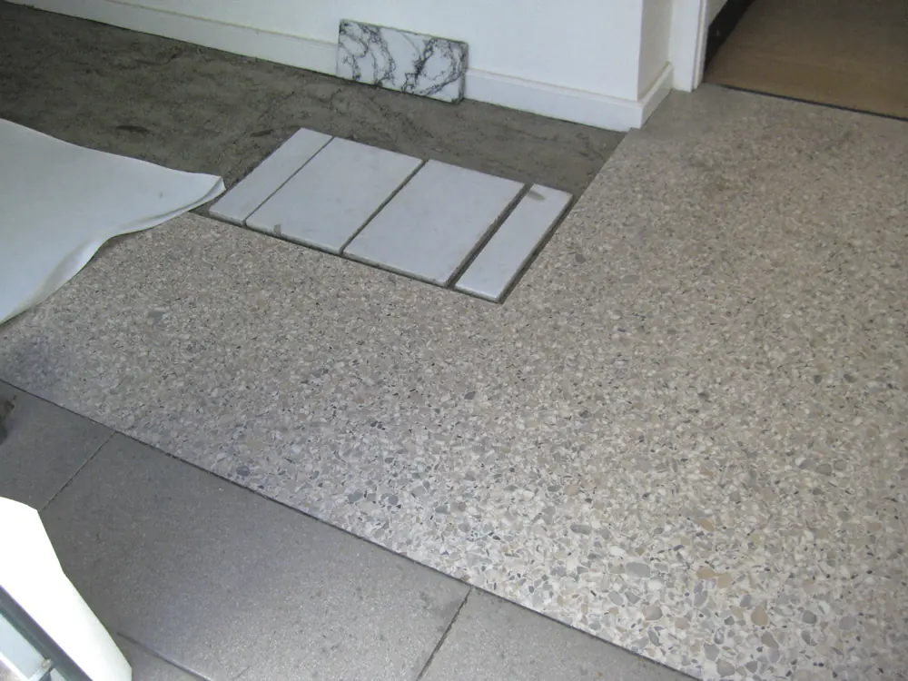 Een oude terrazzovloer in een hal waar marmeren platen zijn losgemaakt, met het mortelbed zichtbaar